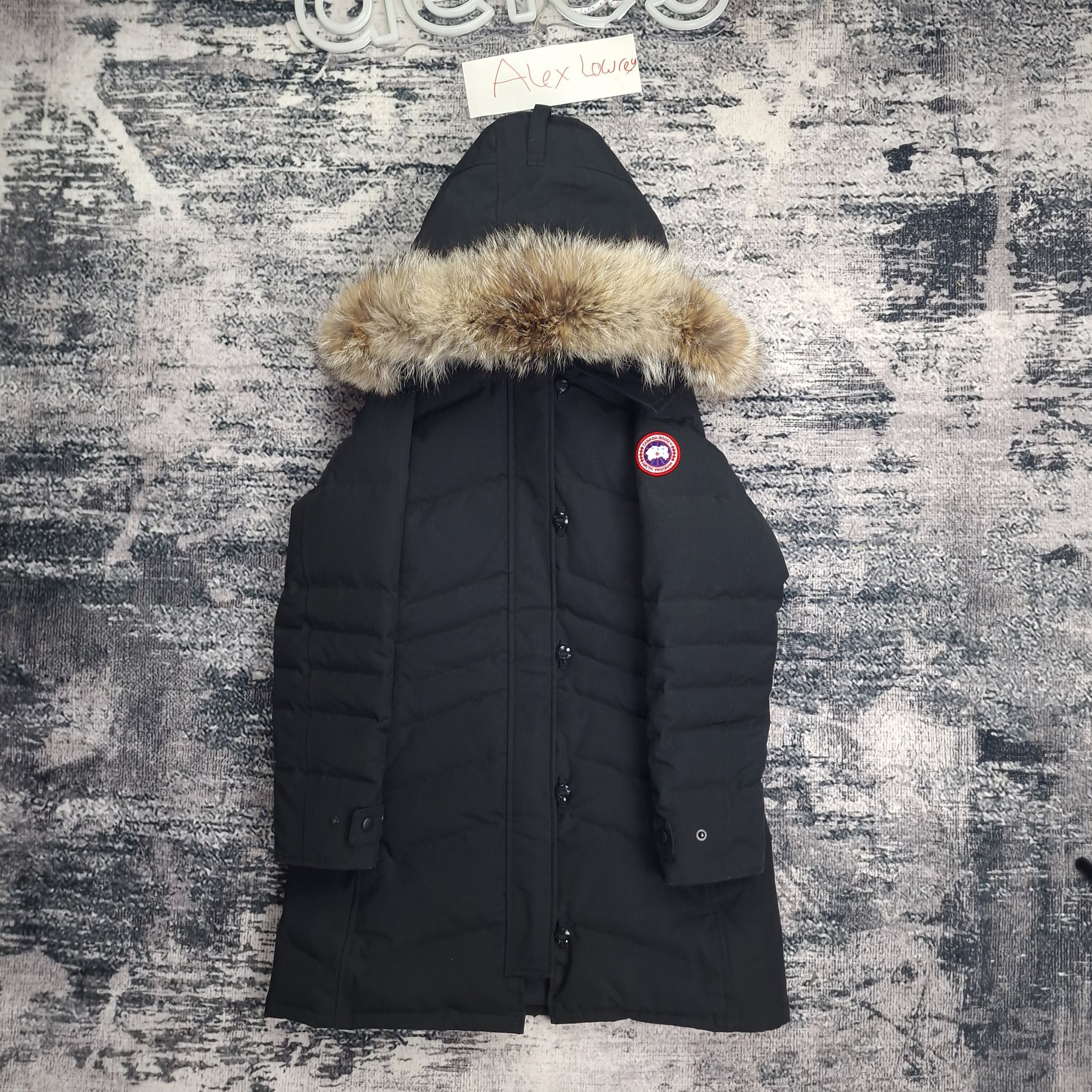 Front flat lay of black Canada Goose Lorette Parka women's medium with fur-trimmed hood, Arctic badge, and elegant knee-length design